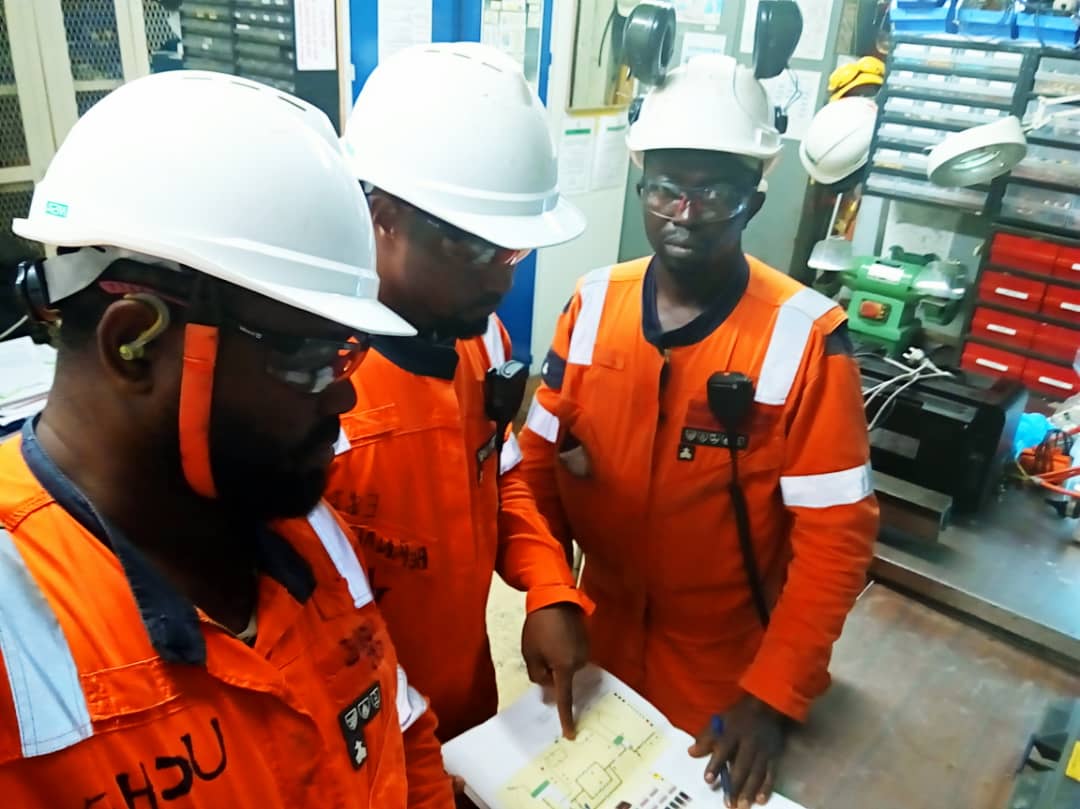 Three technicians in orange PPE are collaborating in a workshop to review a technical schematic diagram
